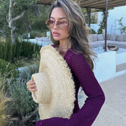 Donna con occhiali da sole e cappello di paglia, indossa abito viola, in giardino mediterraneo. Moda estiva, stile elegante, look casual chic.