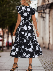 Abito lungo nero a fiori bianchi, donna di spalle, strada cittadina, sandali con tacco, look estivo elegante, moda femminile casual.