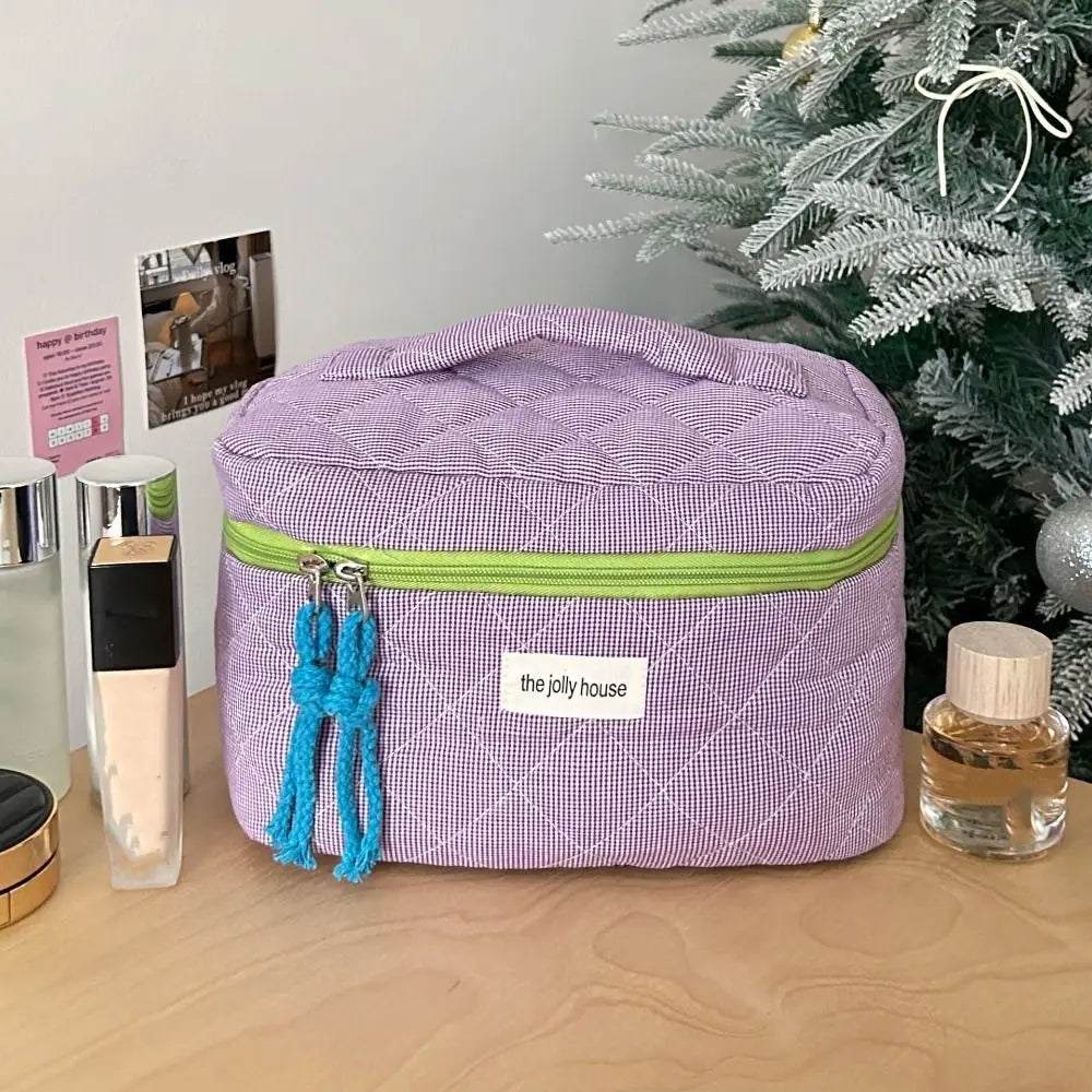 Astuccio cosmetico viola con cerniera verde e nappine blu, su tavolo vicino ad albero di Natale. Accessori trucco e profumo visibili.