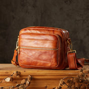 Borsa a tracolla in pelle marrone su tavolo di legno, design elegante con cerniere dorate, ideale per moda uomo e donna, accessorio di tendenza.