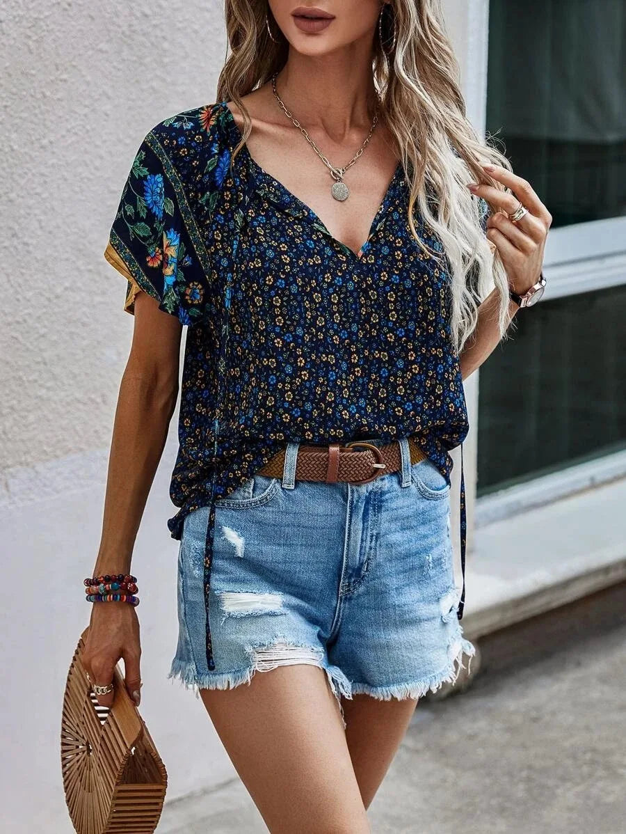 Donna con top floreale blu e shorts di jeans strappati, accessoriata con borsa in legno e bracciali colorati. Moda estiva casual e chic.