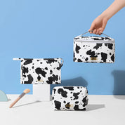 Set di tre beauty case con stampa mucca bianca e nera su sfondo blu. Include pennello trucco. Design moderno, accessori moda eleganti.