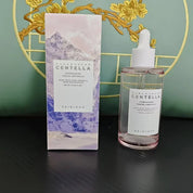 Boccetta di siero viso Skin1004 Madagascar Centella, 100 ml, con scatola decorata da montagne e fiori gialli, su sfondo verde.
