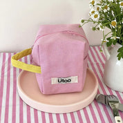 Astuccio rosa in tessuto con manico giallo su piatto rosa, decorato con fiori bianchi e verdi. Accessorio moda, design elegante e funzionale.