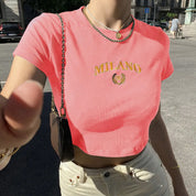 Donna con t-shirt rosa con scritta "Milano", collana dorata, borsa a tracolla nera, jeans chiari. Moda estiva urbana, stile casual chic.