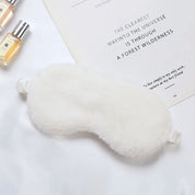 Maschera per dormire in pelliccia bianca su sfondo bianco, accanto a flaconi di profumo e un libro. Accessorio relax, comfort, sonno riposante.