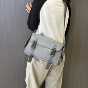 Borsa a tracolla grigia unisex, design moderno, chiusure a fibbia, indossata su maglia bianca e nera, pantaloni beige. Accessorio moda casual.