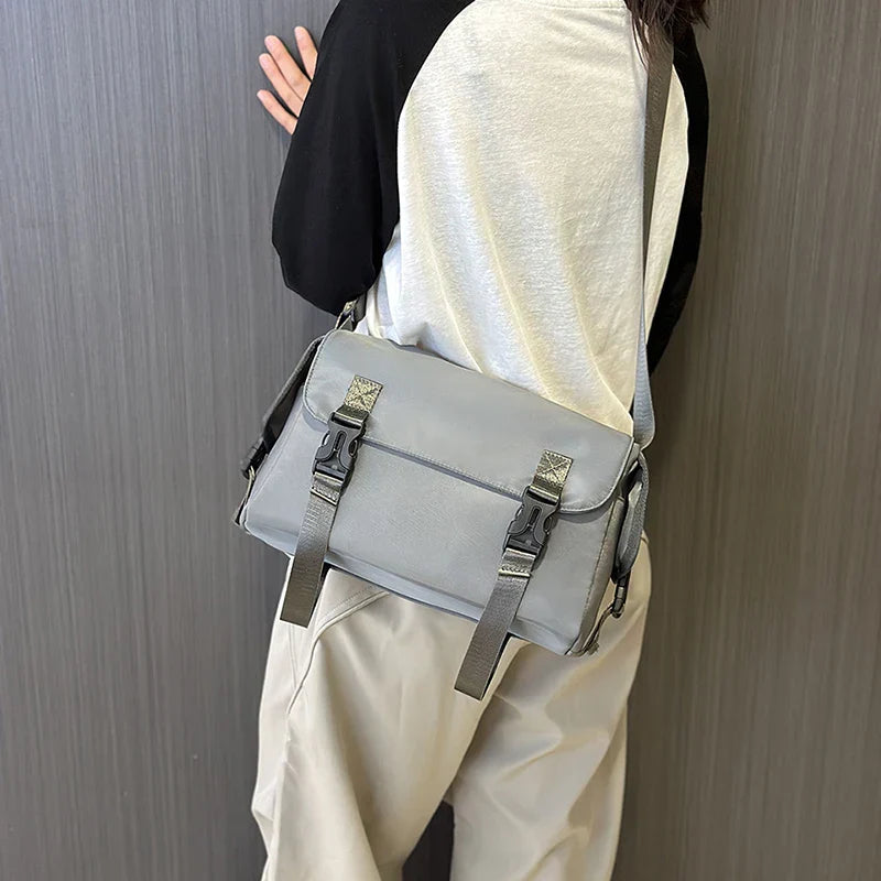Borsa a tracolla grigia unisex, design moderno, chiusure a fibbia, indossata su maglia bianca e nera, pantaloni beige. Accessorio moda casual.