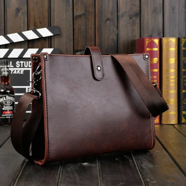 Borsa a tracolla in pelle marrone su tavolo di legno, con sfondo di libri e bottiglia. Elegante accessorio moda uomo, stile vintage.