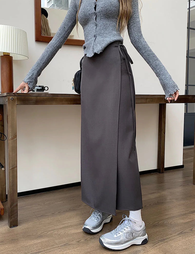 Donna in maglione grigio e gonna lunga marrone, scarpe da ginnastica argentate, in piedi accanto a un tavolo di legno. Outfit casual autunnale.