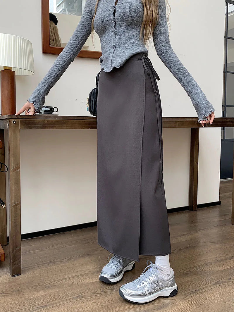 Donna in maglione grigio e gonna lunga marrone, scarpe da ginnastica argentate, in piedi accanto a un tavolo di legno. Outfit casual autunnale.