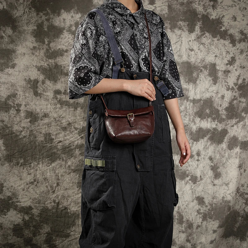 Persona con tuta nera e camicia a motivi geometrici, indossa una borsa a tracolla in pelle marrone. Stile casual, moda urbana, accessori vintage.