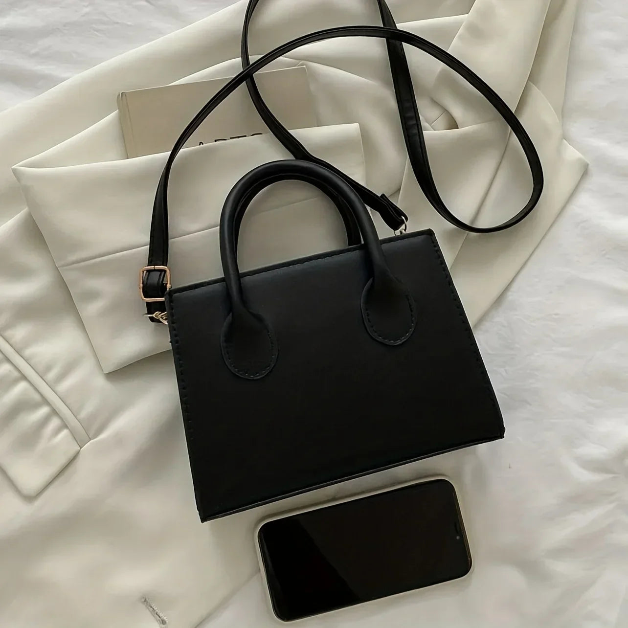 Borsa a mano nera elegante su giacca bianca, con smartphone e libro. Accessorio moda donna, stile minimalista, pelle sintetica.