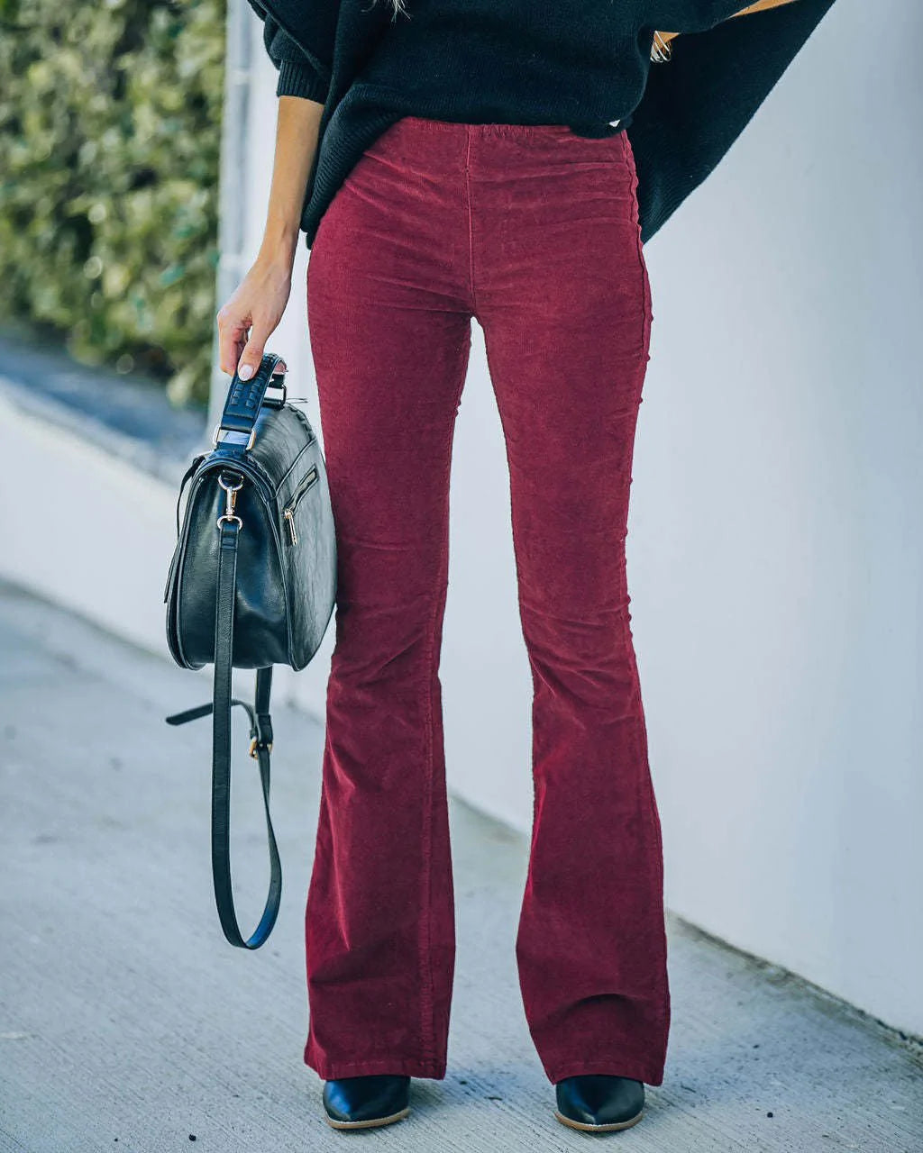 Donna con pantaloni a zampa bordeaux, maglione nero, borsa a tracolla nera e stivali neri. Moda autunnale, abbigliamento casual elegante.