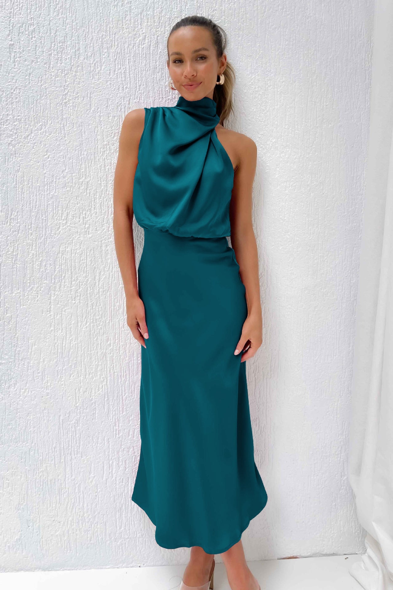 Abito lungo elegante verde smeraldo con collo alto drappeggiato, indossato da una donna sorridente su sfondo bianco. Moda femminile sofisticata.