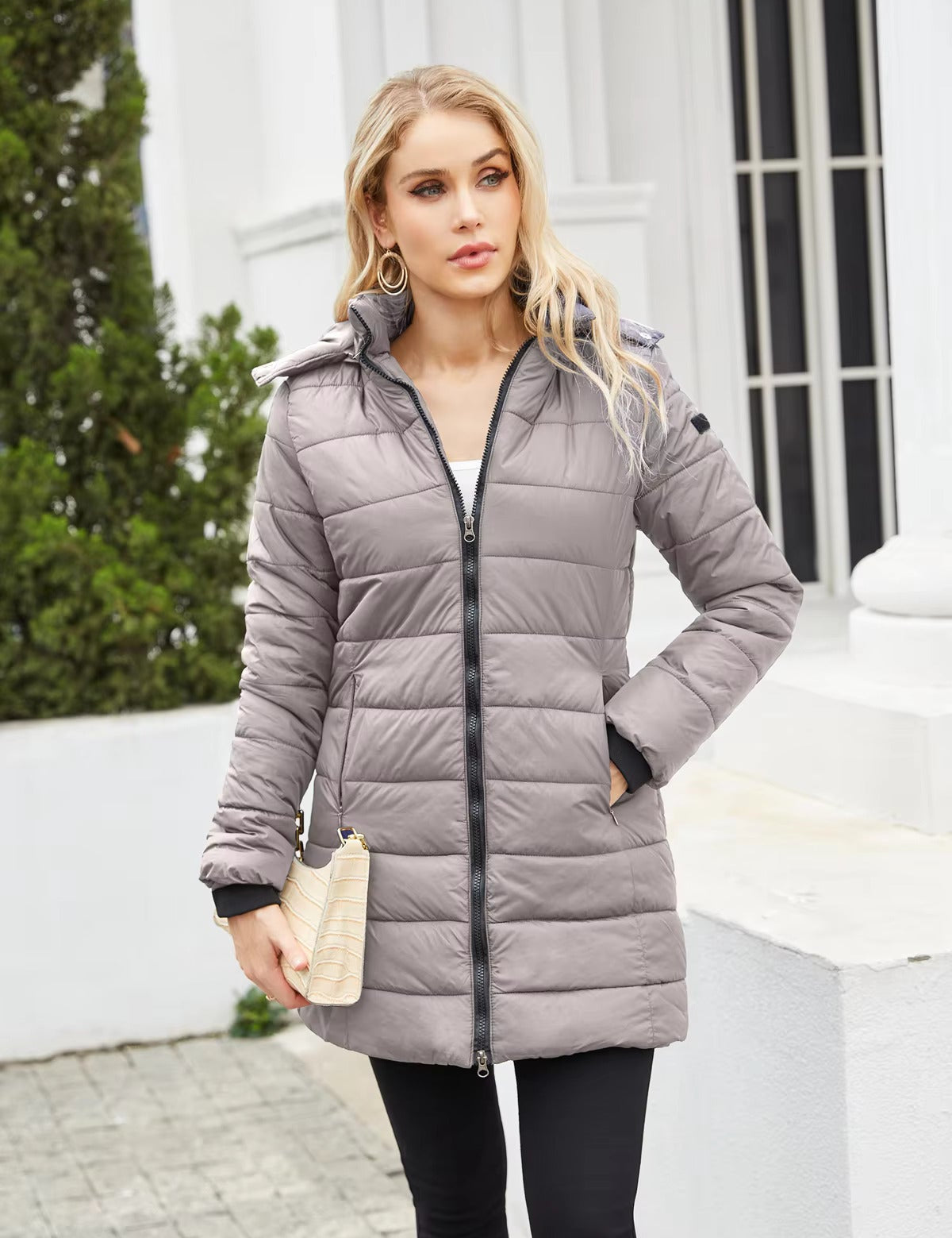 Donna con cappotto imbottito grigio, zip frontale, cappuccio, tasche laterali, posa all'aperto. Moda invernale, abbigliamento caldo, stile casual.