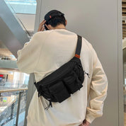 Uomo con marsupio nero a tracolla in centro commerciale moderno, indossa felpa beige e cappellino nero. Accessorio moda casual, pratico e spazioso.