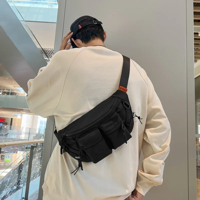 Uomo con marsupio nero a tracolla in centro commerciale moderno, indossa felpa beige e cappellino nero. Accessorio moda casual, pratico e spazioso.