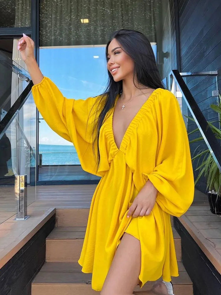 Donna con abito giallo estivo, scollo a V profondo, maniche ampie, su terrazza moderna con vista mare. Moda estiva, abbigliamento femminile elegante.