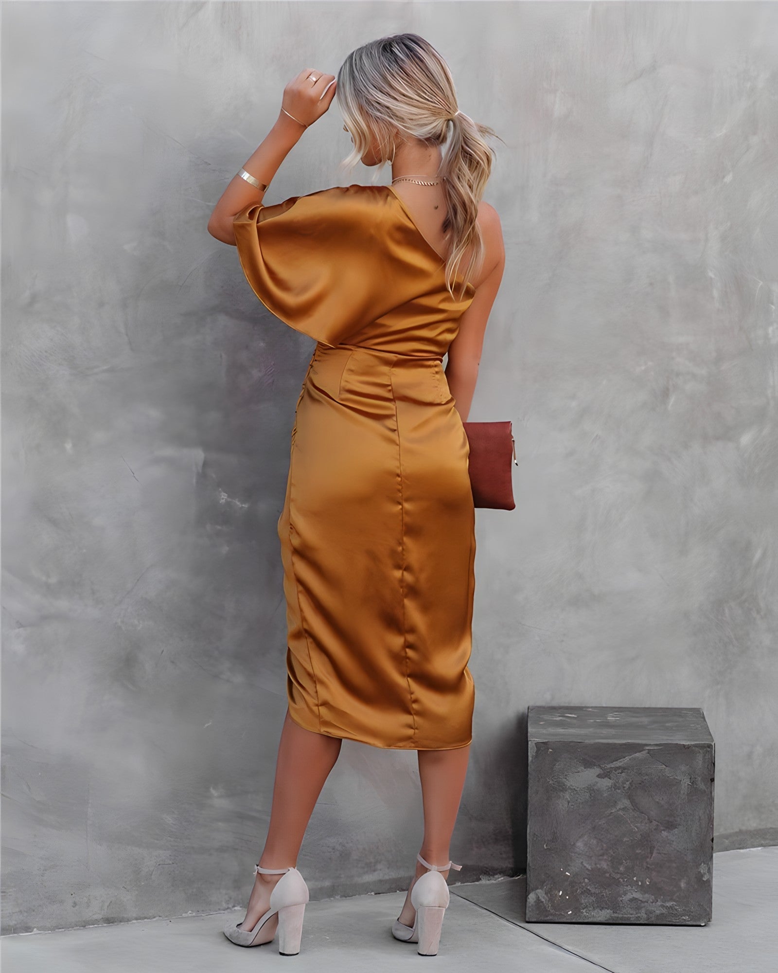Donna in abito satinato color oro, spalle scoperte, con pochette marrone e tacchi alti, posa contro parete grigia. Moda elegante e sofisticata.