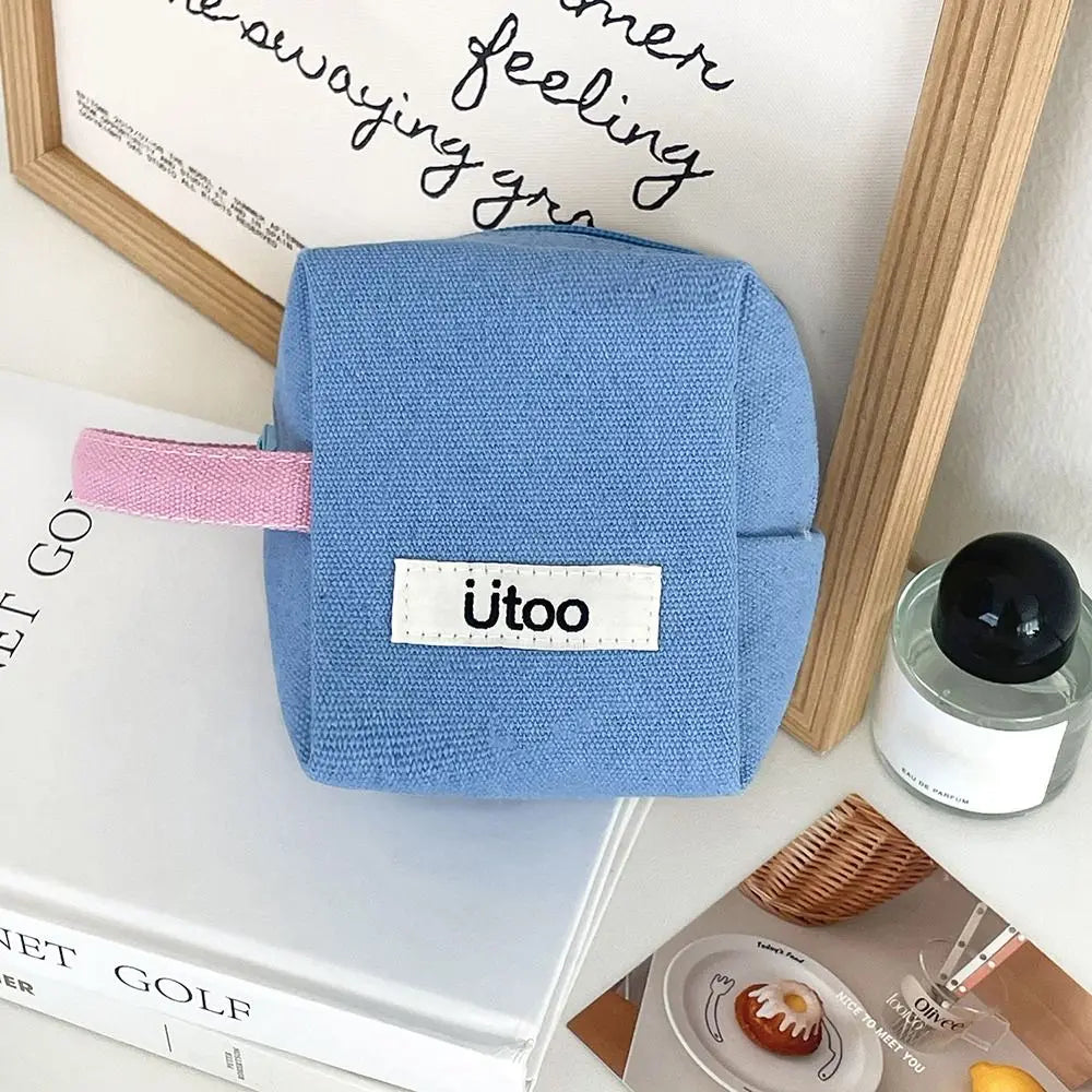 Astuccio quadrato in tessuto blu con etichetta "ütoo", manico rosa, su tavolo con cornice in legno, libro e candela. Accessorio scuola, organizer.