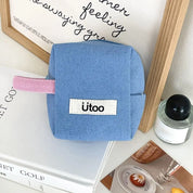 Astuccio quadrato in tessuto blu con etichetta "ütoo", manico rosa, su tavolo con cornice in legno, libro e candela. Accessorio scuola, organizer.