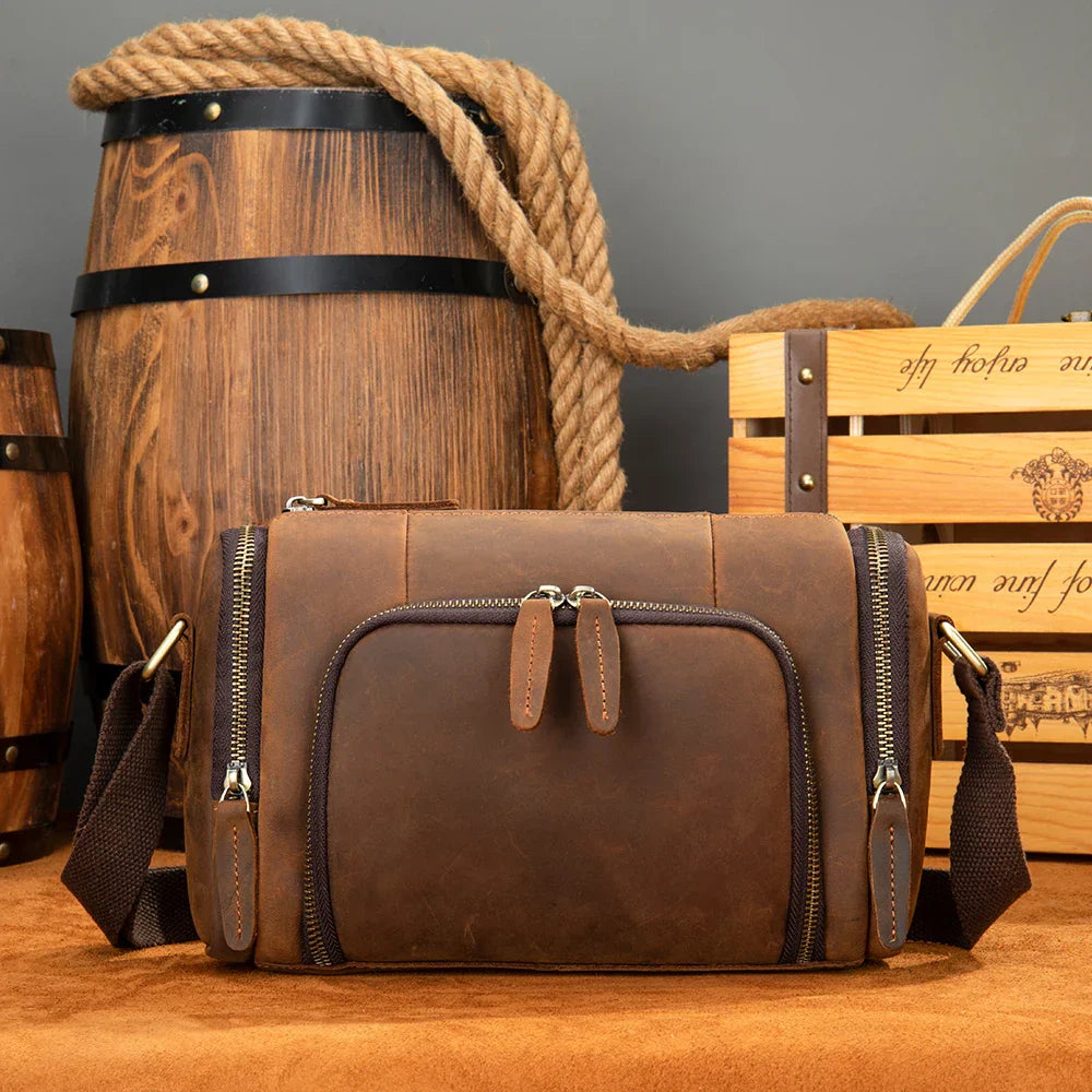 Borsa a tracolla in pelle marrone vintage, con cerniere multiple, davanti a barili di legno e cassa di vino. Accessorio moda uomo elegante e pratico.