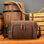 Borsa a tracolla in pelle marrone vintage, con cerniere multiple, davanti a barili di legno e cassa di vino. Accessorio moda uomo elegante e pratico.