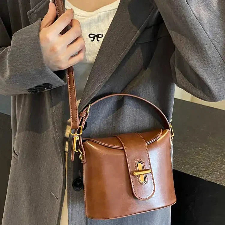 Borsa a tracolla in pelle marrone, design elegante con chiusura a fibbia dorata, indossata su cappotto grigio. Accessorio moda donna.