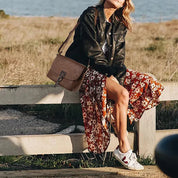 Donna seduta su una staccionata, indossa giacca di pelle nera, gonna floreale rossa e sneakers bianche, con borsa a tracolla marrone.