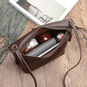 Borsa a tracolla in pelle marrone aperta su tavolo di legno, contenente smartphone, portafoglio, profumo e rossetto. Accessori moda eleganti.