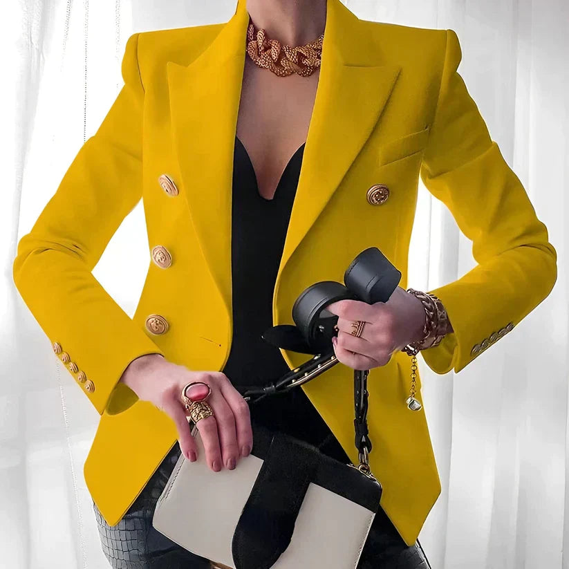 Giacca gialla elegante con bottoni dorati, indossata sopra top nero. Accessori: collana dorata, borsa bianca e nera, anelli e bracciale.