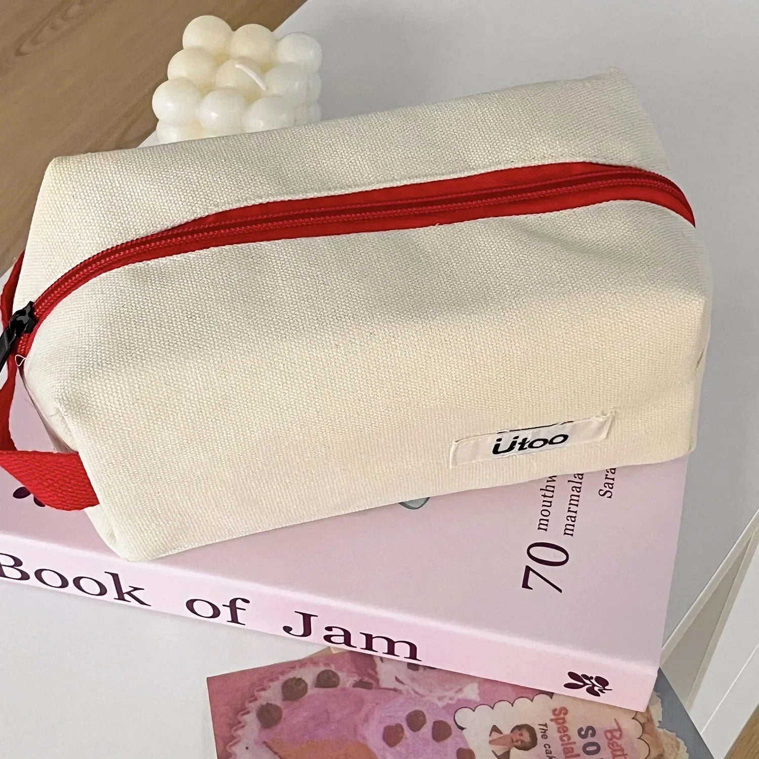 Borsa da toilette in tessuto beige con cerniera rossa, posizionata su un libro rosa. Design moderno, ideale per viaggi e organizzazione.