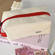 Borsa da toilette in tessuto beige con cerniera rossa, posata su un libro rosa. Design moderno, ideale per viaggi e organizzazione accessori.