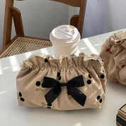 Borsa beige con fiocco nero e motivi a pois neri, accanto a bicchiere di caffè su tavolo. Accessorio moda elegante, perfetto per ogni occasione.