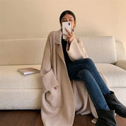 Donna seduta su divano bianco, indossa cappotto beige oversize, jeans blu e stivali neri. Smartphone in mano, libro accanto. Moda casual autunnale.