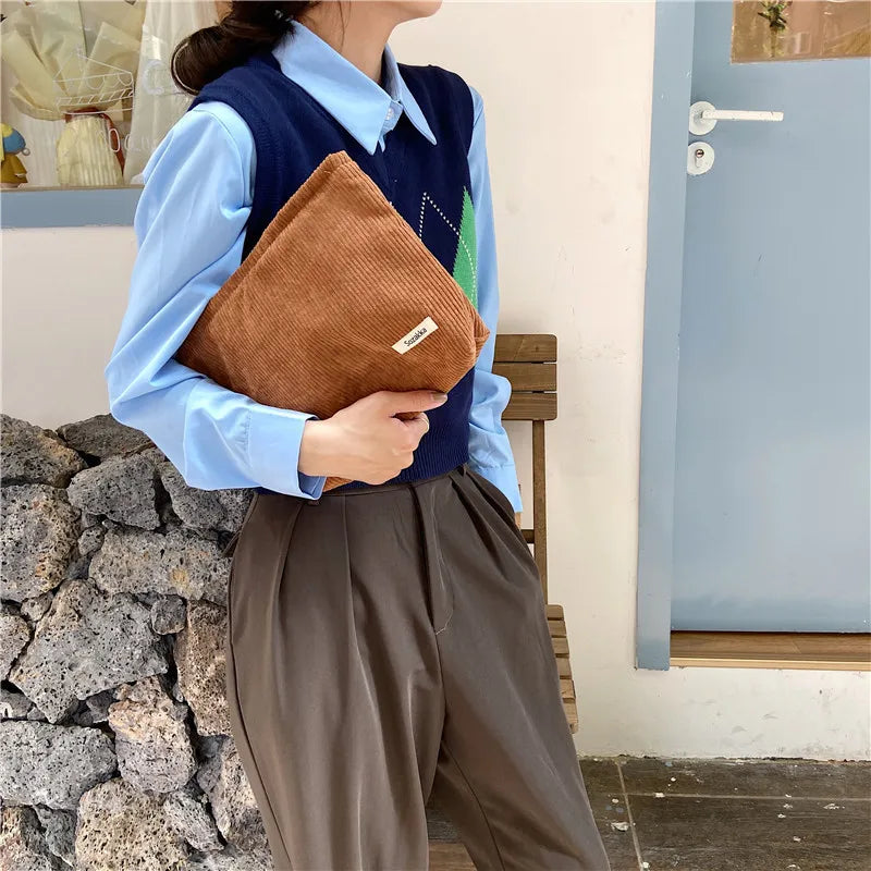 Donna con camicia azzurra e gilet blu, tiene una borsa a mano marrone in velluto a coste. Moda autunnale, abbigliamento casual elegante.