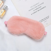 Mascherina per dormire in peluche rosa su sfondo bianco, accanto a flaconi di profumo e libro. Accessorio per il sonno, morbido e confortevole.