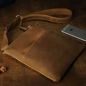 Borsa a tracolla in pelle marrone su tavolo di legno, design elegante e minimalista, ideale per uomo, con smartphone accanto.