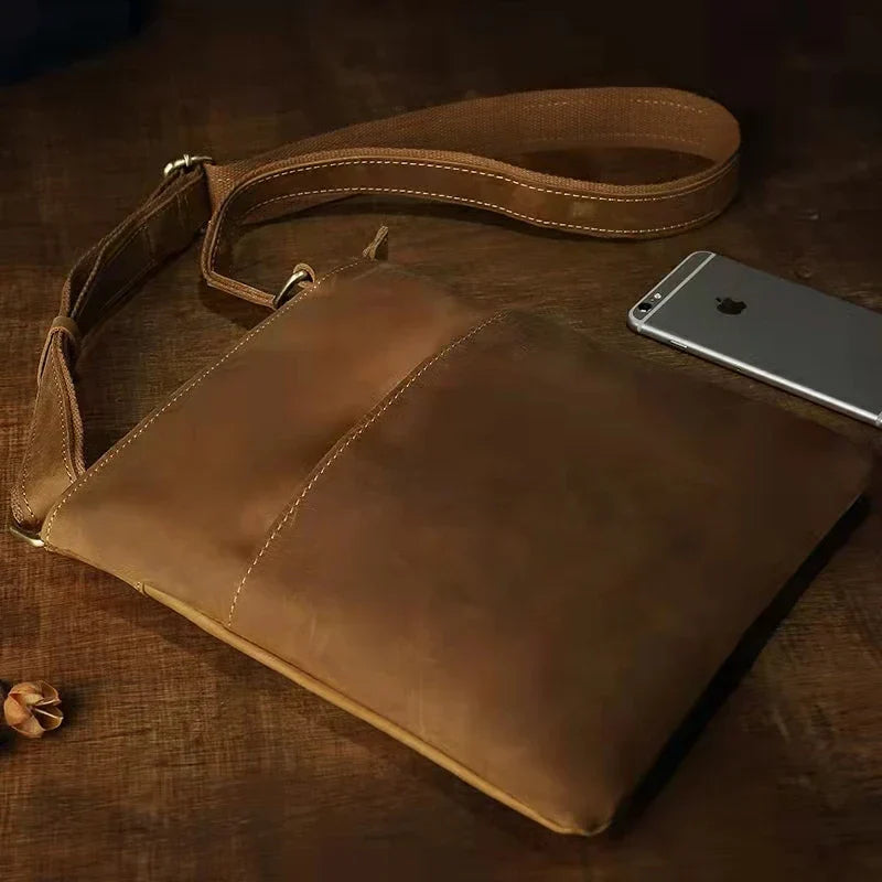 Borsa a tracolla in pelle marrone su tavolo di legno, design elegante e minimalista, ideale per uomo, con smartphone accanto.