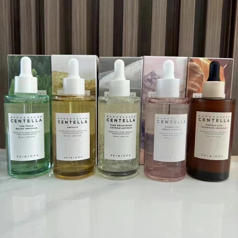 Cinque flaconi di siero SKIN1004 Centella su sfondo a righe, con diverse formule per la cura della pelle. Prodotti skincare coreani.