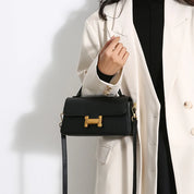 Donna con cappotto bianco indossa borsa a tracolla nera elegante con chiusura dorata. Accessorio moda femminile, stile chic e moderno.