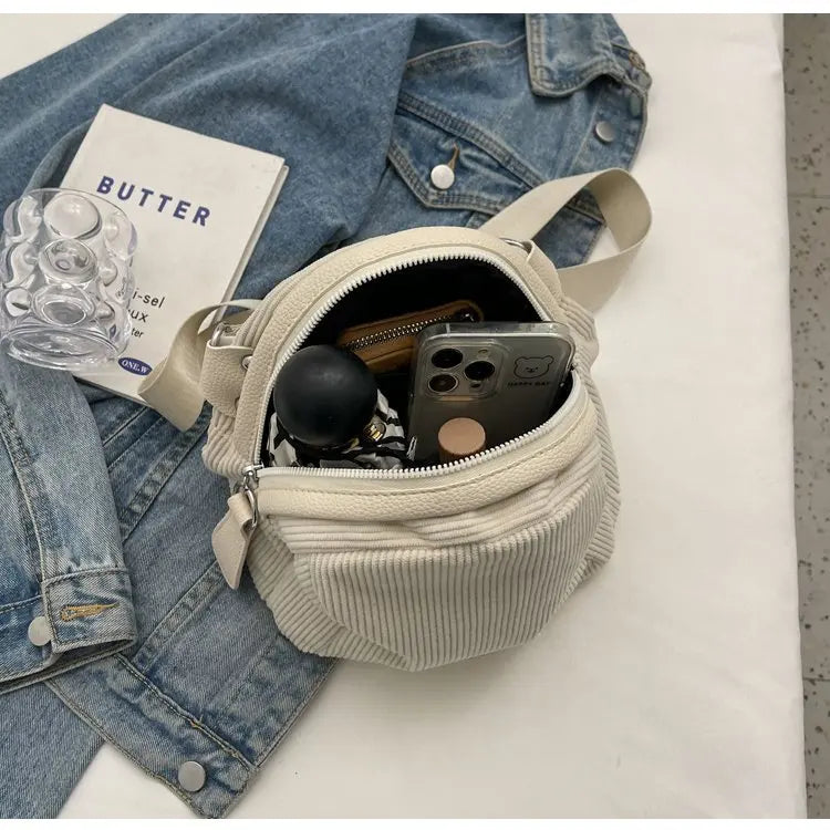 Borsa a tracolla beige in tessuto a coste, contenente smartphone, portafoglio e accessori, su giacca di jeans e libro. Accessori moda casual.