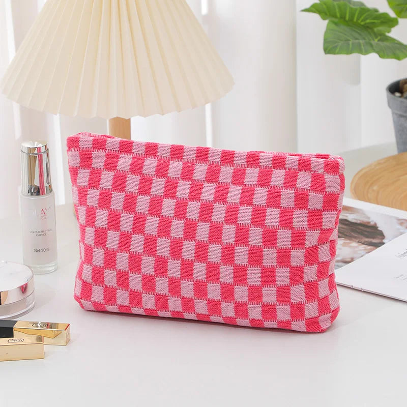 Astuccio rosa a quadretti bianchi su tavolo, accanto a lampada e pianta. Design moderno, ideale per cosmetici o accessori.