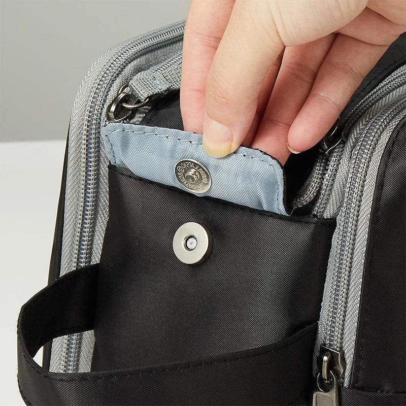 Mano che apre tasca con bottone a pressione su zaino nero in tessuto resistente, cerniere grigie. Accessorio pratico per viaggio e lavoro.