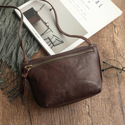 Borsa a tracolla in pelle marrone su tavolo di legno, accanto a occhiali e rivista. Design elegante, chiusura con zip, accessorio moda donna.