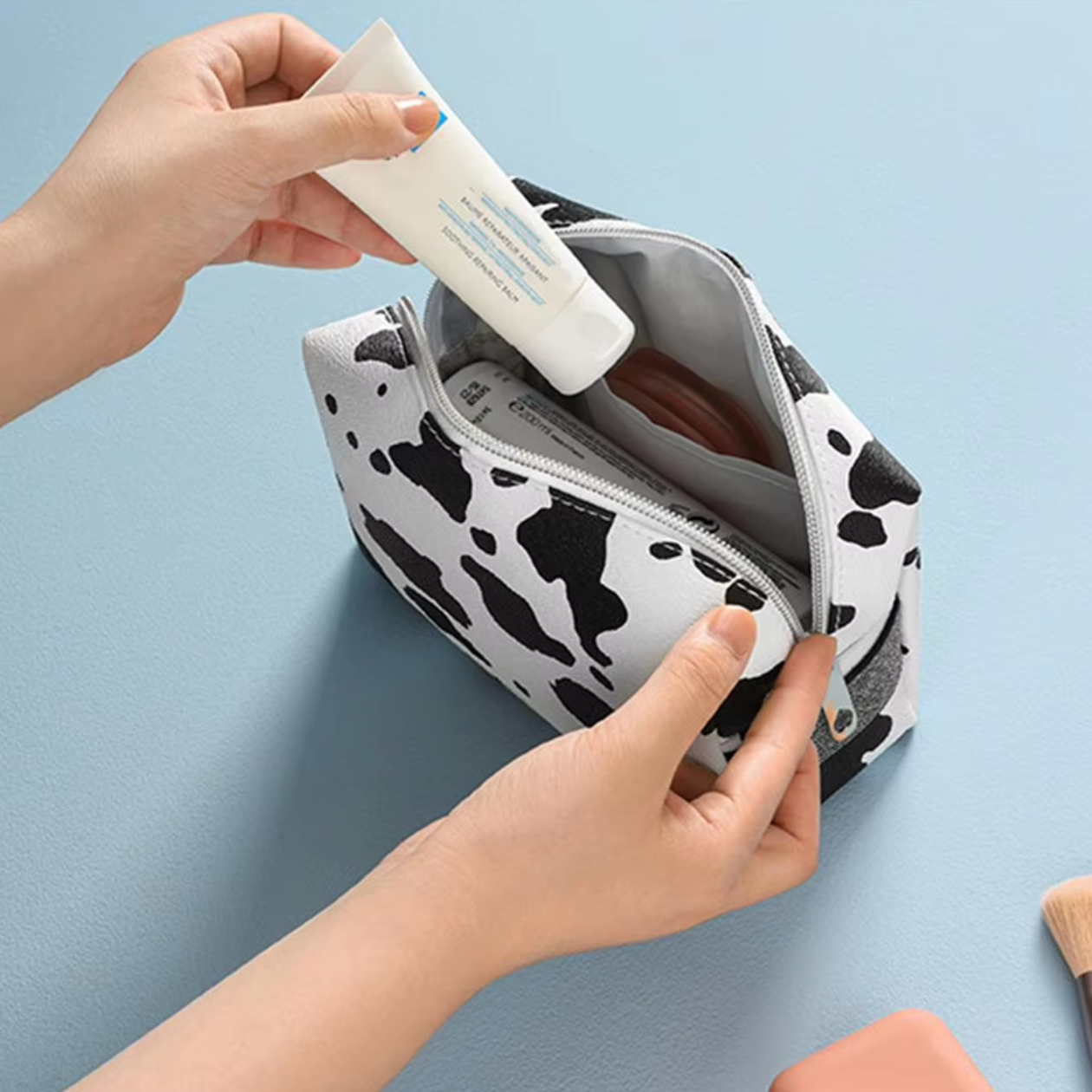 Mani che inseriscono un tubetto di crema in un astuccio cosmetico bianco e nero a macchie su sfondo azzurro. Accessori trucco visibili.