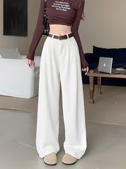 Donna con pantaloni bianchi larghi, cintura marrone e top marrone a maniche lunghe. Stile moda casual, interno moderno, laptop su tavolo.