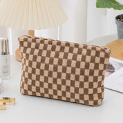Borsa a scacchi marrone e beige su tavolo bianco, accanto a lampada e piante. Design elegante, ideale per moda e accessori.