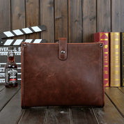 Cartella in pelle marrone su sfondo di legno, con bottiglia di whisky e libri. Elegante e resistente, ideale per lavoro e ufficio.